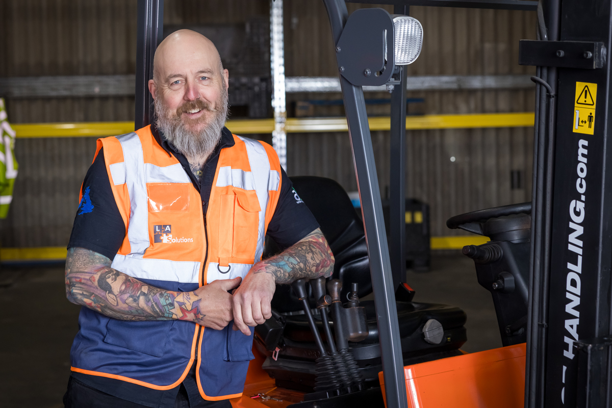picture of a tattooed man called Lance standing next to a orange forklift